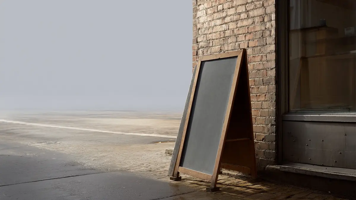A blank chalkboard sign leaning against a brick shopfront on a quiet damp pavement in early morning light.