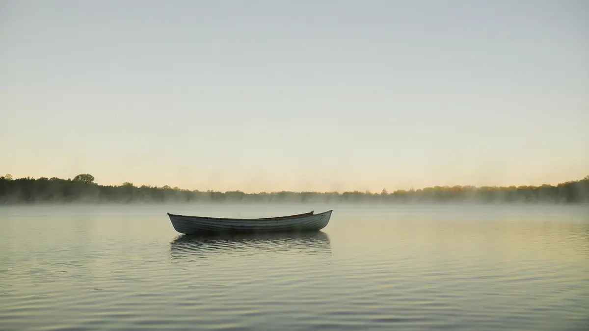 A wooden rowing boat resting on still misty water at dawn with soft pale light coming through distant trees.