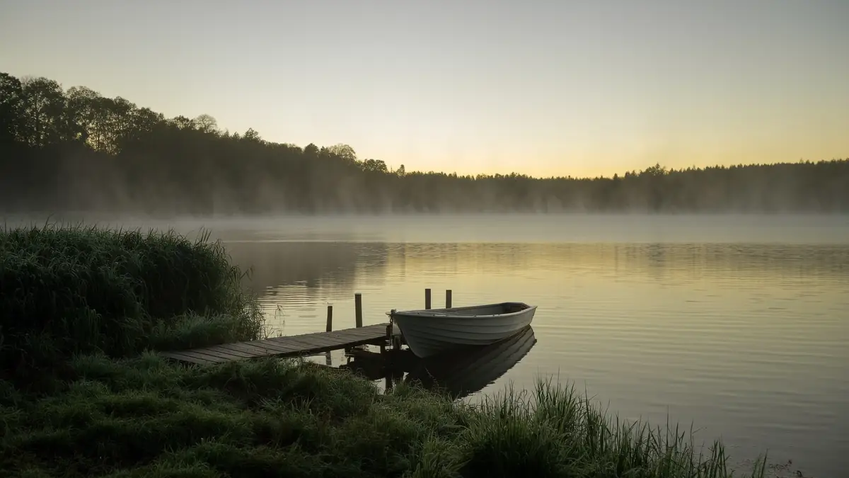 A wooden rowing boat tied to a quiet lakeside jetty in soft early morning mist.