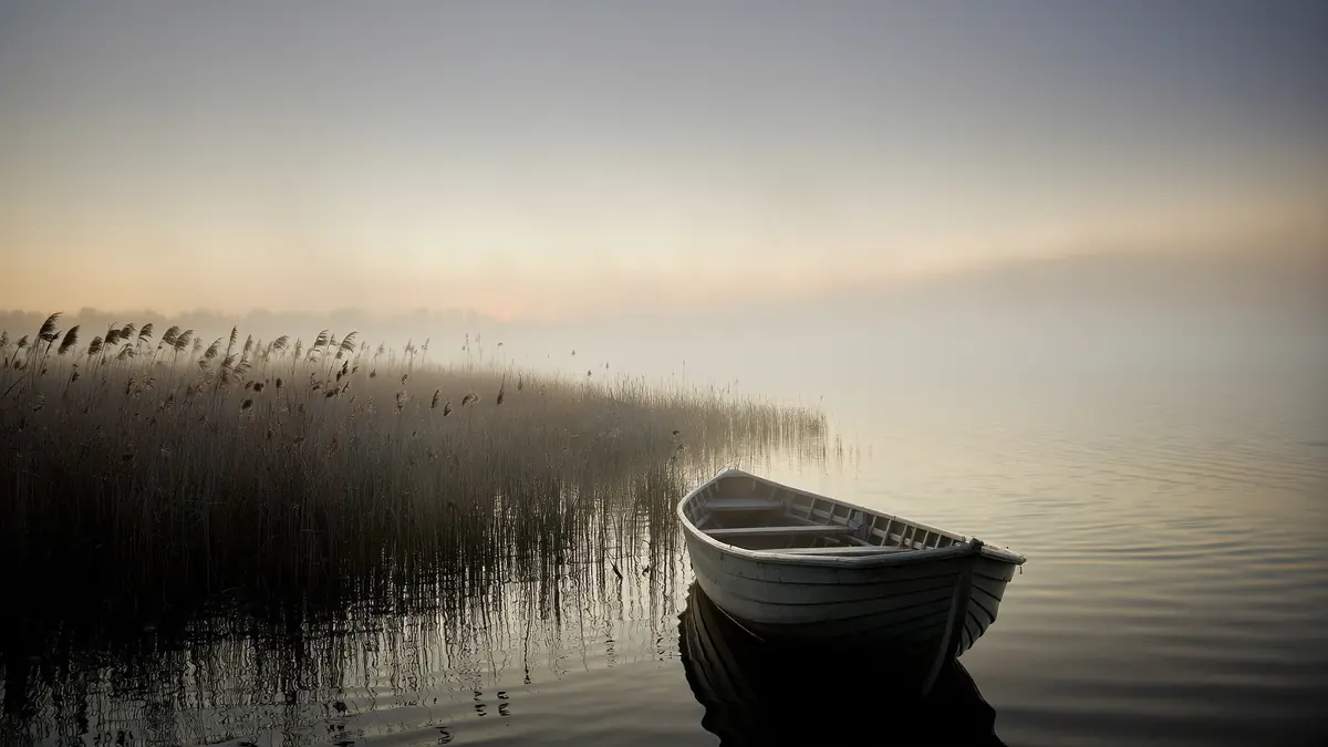 A wooden rowing boat moored at a misty lakeside at dawn with soft pale light reflected in still water.
